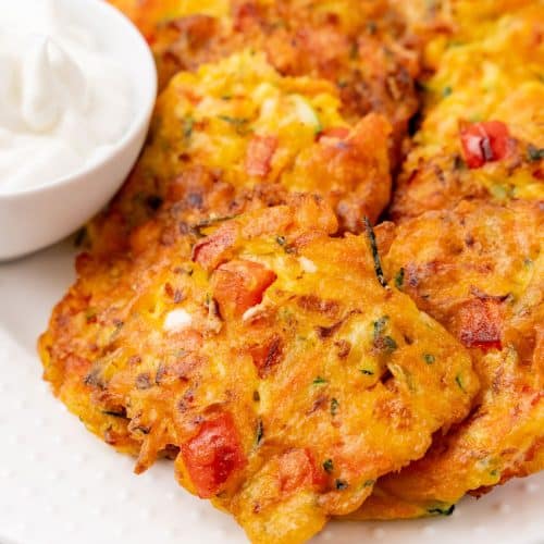 carrot and zucchini fritters with feta piled on a white serving platter with a small bowl of plain yogurt on the side