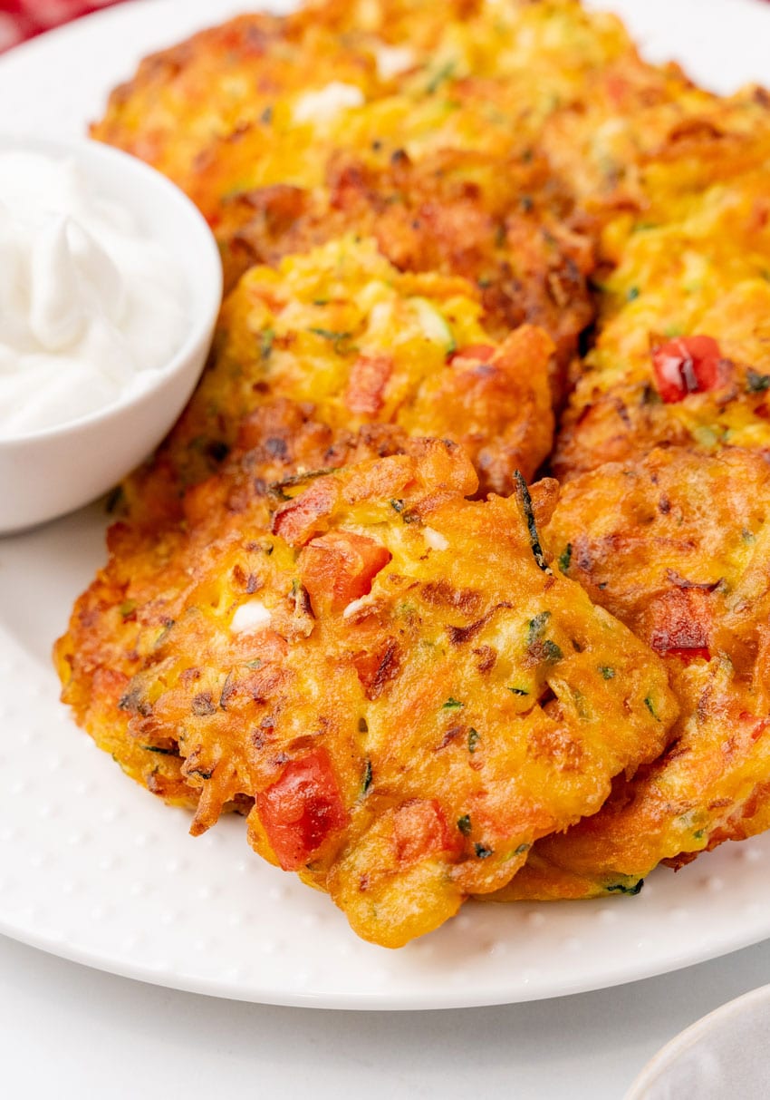 carrot and zucchini fritters with feta piled on a white serving platter with a small bowl of plain yogurt on the side