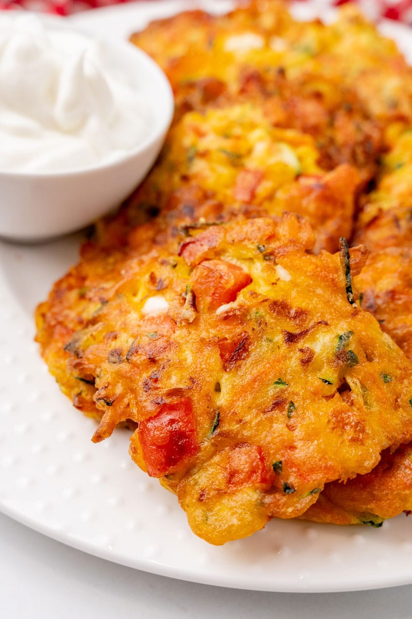 carrot and zucchini fritters with feta piled on a white serving platter with a small bowl of plain yogurt on the side