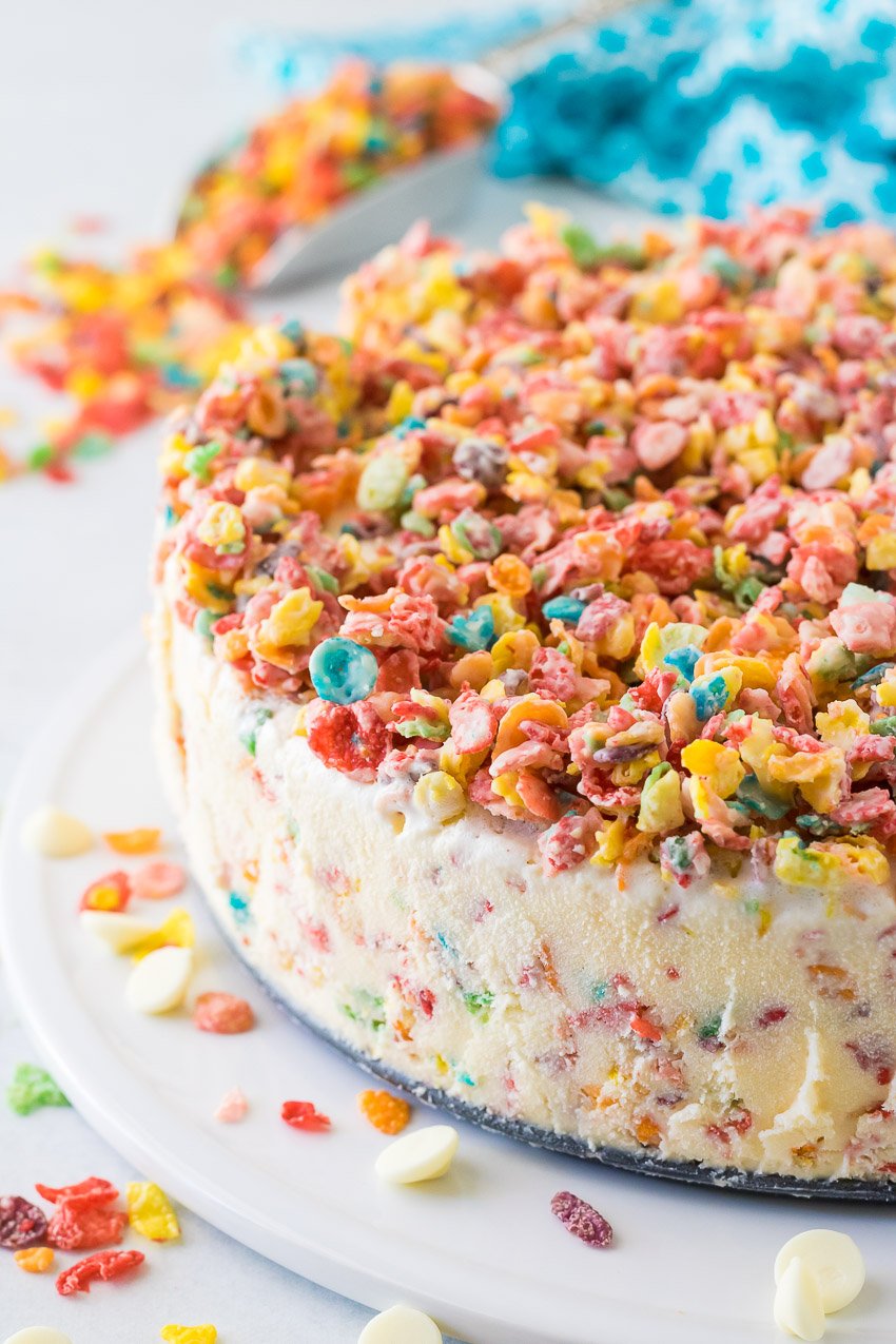 fruity pebble ice cream cake on a white serving tray surrounded by colorful breakfast cereal pieces