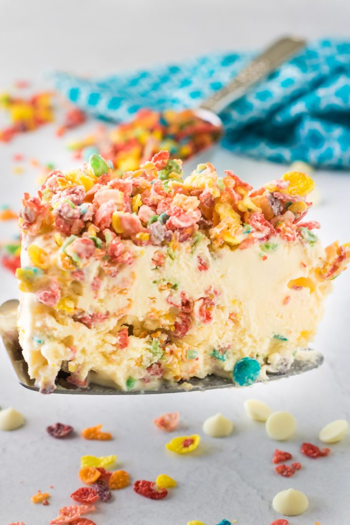 a colorful slice of Fruity Pebble Ice Cream Cake is held up on a metal cake server with a bright teal blue cloth in the background and a scoop of fruity pebbles spilling over around it