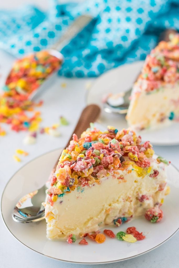 two slices of this creamy Fruity Pebble Ice Cream Cake served on crisp white porcelain dessert plates with silver spoons on the side