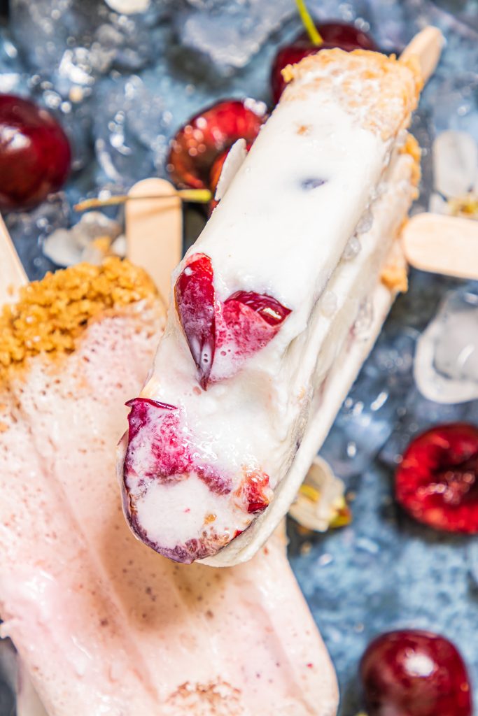 a cherry cheesecake popsicle shown with a bite removed to reveal the sweet dark cherries studding the inside