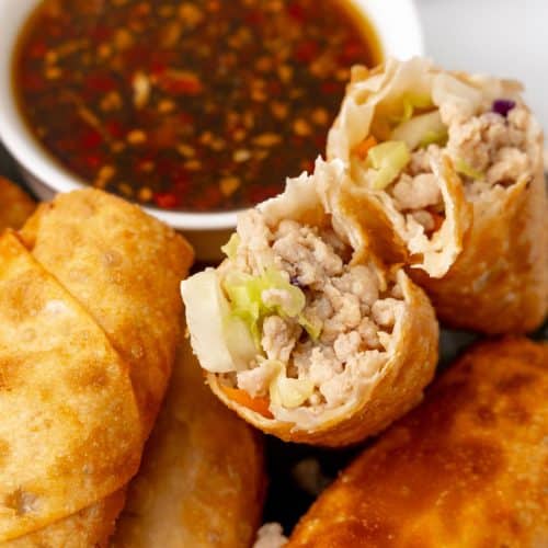 a homemade egg roll broken in half and resting on a plate of egg rolls with a small white bowl filled with sesame garlic dipping sauce on the side