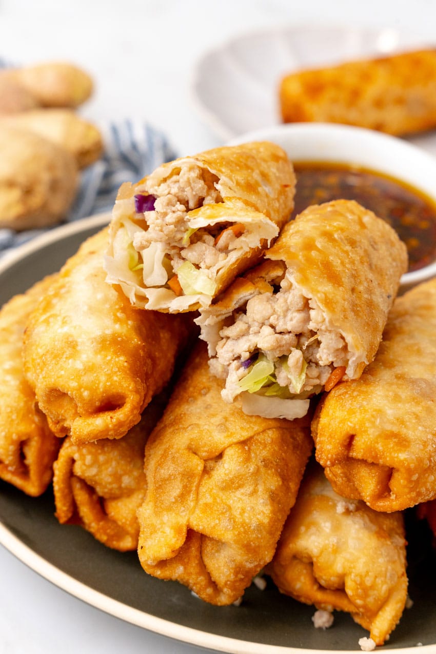 a homemade egg roll broken in half and resting on a plate of egg rolls with a small white bowl filled with sesame garlic dipping sauce on the side