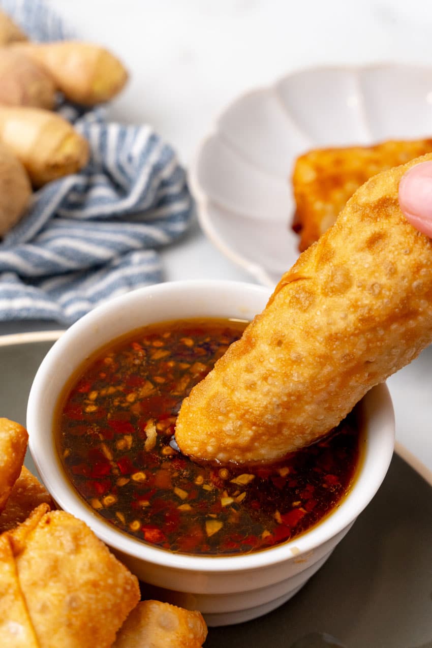 a hand dipping a homemade pork egg roll in a small bowl of sesame ginger dipping sauce