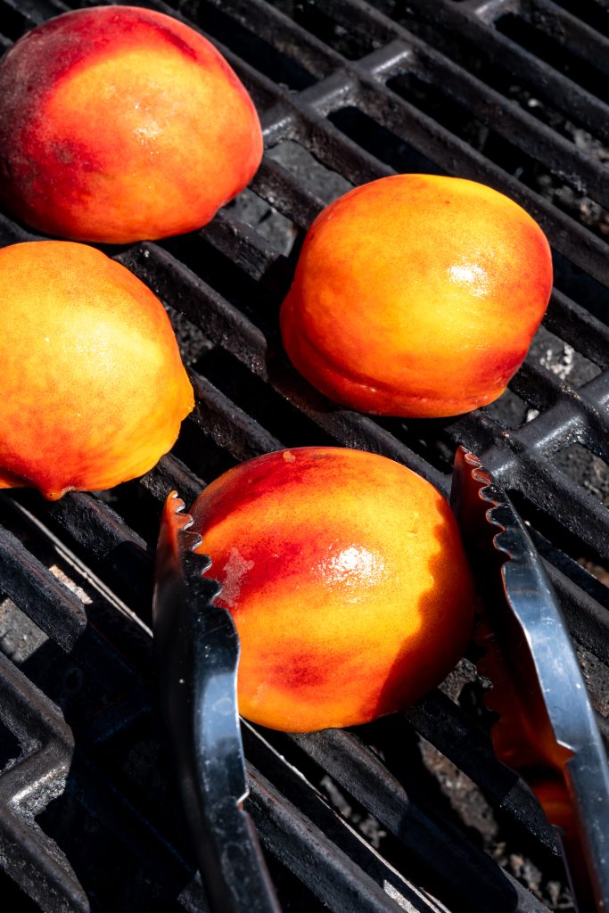 halved peaches on the grates of a grill next to a pair of silver tongs
