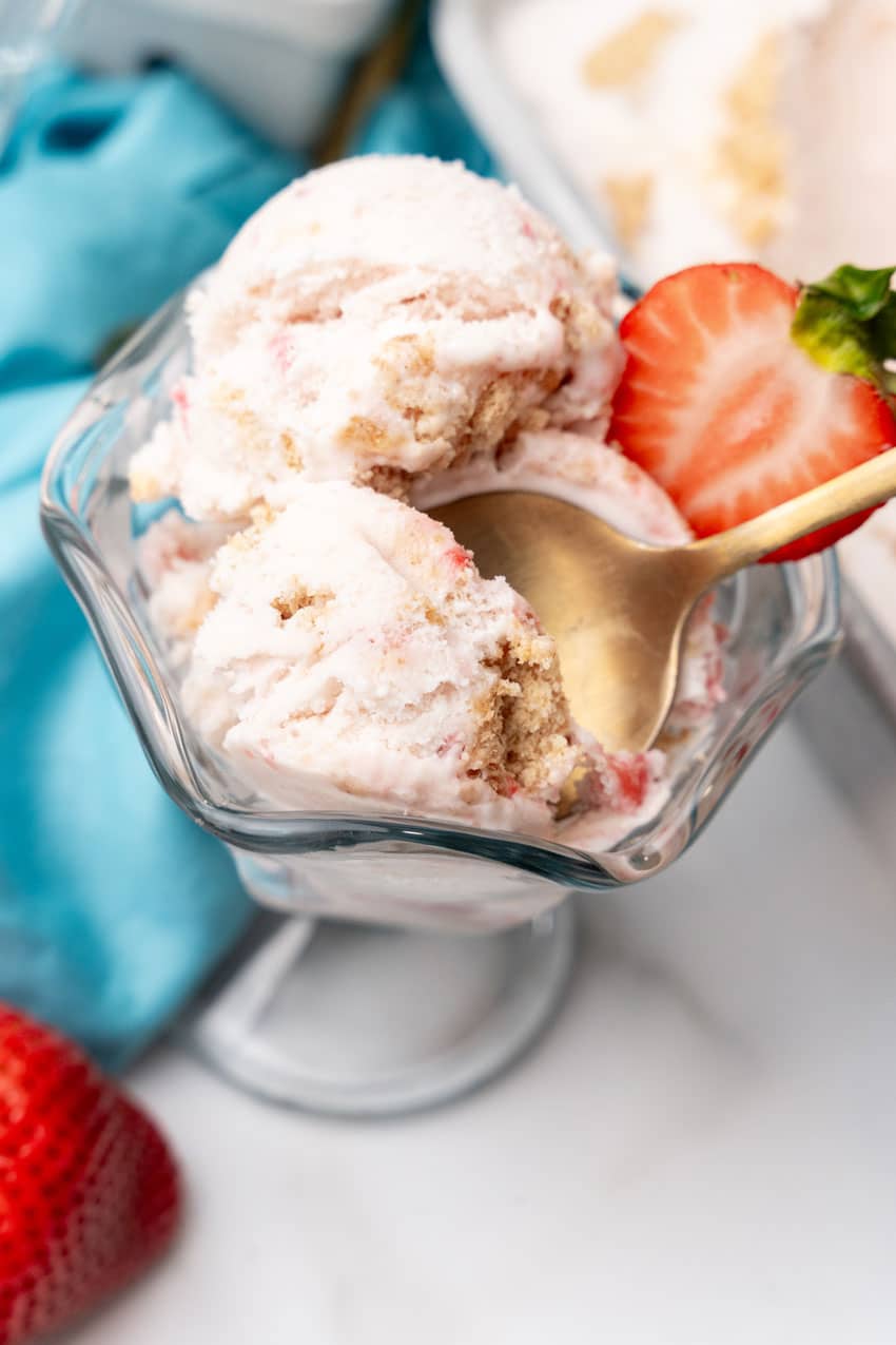 a gold spoon scooping strawberry cheesecake ice cream out of a glass ice cream dish
