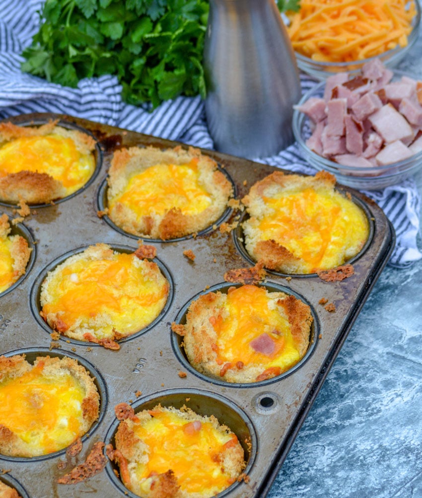 Ham, Egg, & Cheese Toast Cups in the silver muffin tin they were cooked in- shredded cheese, chopped ham, and fresh herbs in the background