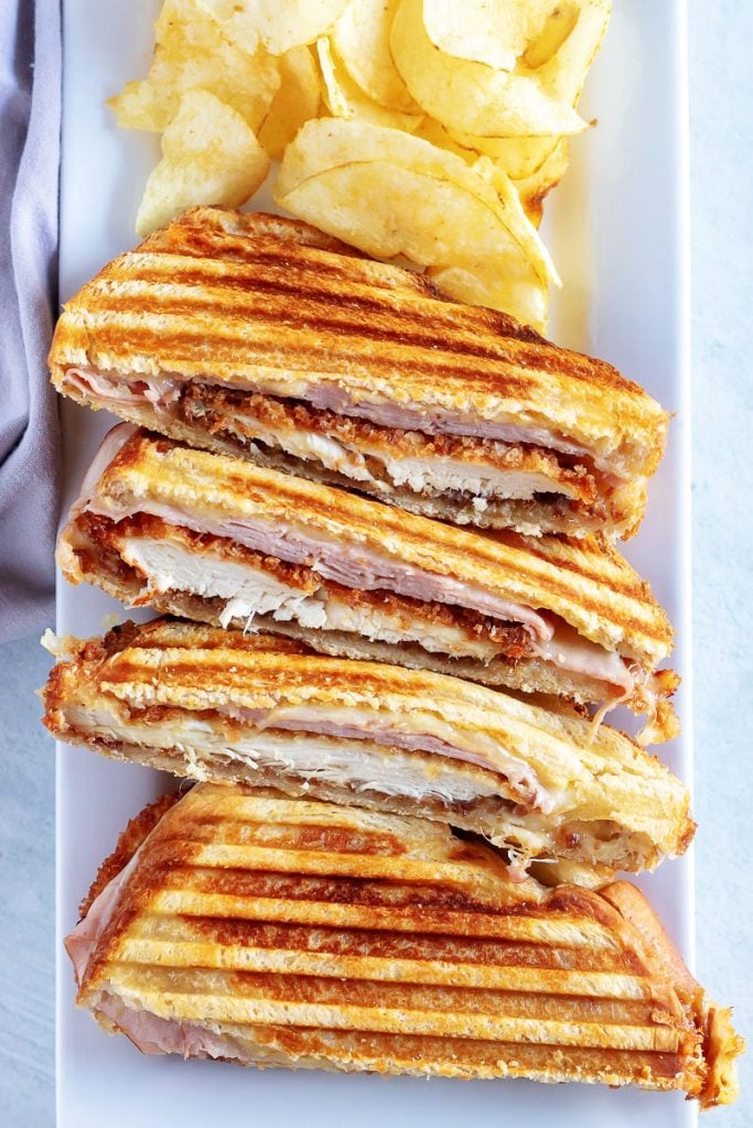 Chicken Cordon Bleu Panini cut in half and displayed on a white serving platter with potato chips to show the crispy chicken cutlets, sliced ham, and melted Swiss