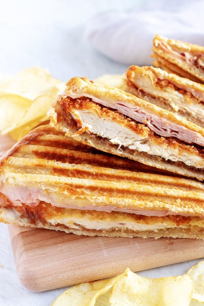 Chicken Cordon Bleu Paninis sliced in half and stacked on top of each other are fanned out on a wooden cutting board with potato chips in the background