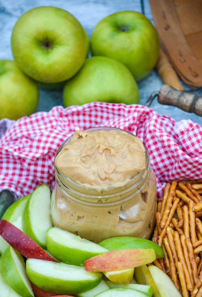 creamy peanut butter apple dip in a small mason jar on a metal serving tray with apples & pretzels with a red & white checkered cloth napkin in the background