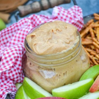 creamy peanut butter apple dip in a small mason jar on a metal serving tray with apples & pretzels with a red & white checkered cloth napkin in the background