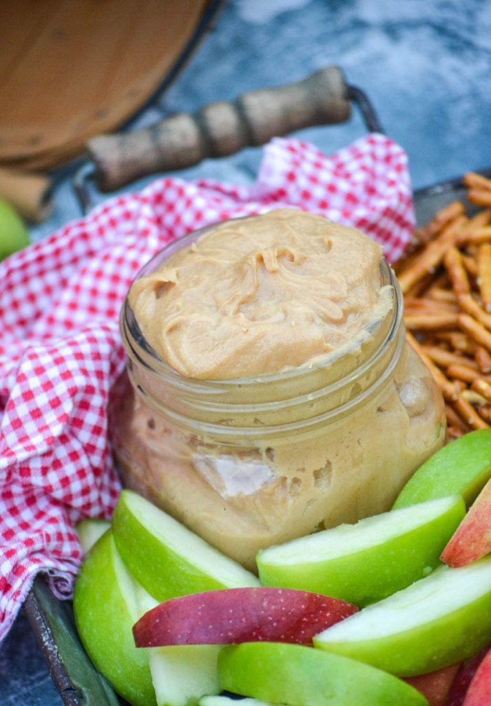 creamy peanut butter apple dip in a small mason jar on a metal serving tray with apples & pretzels with a red & white checkered cloth napkin in the background