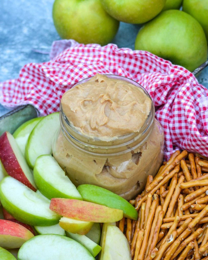 creamy peanut butter apple dip in a small mason jar on a metal serving tray with apples & pretzels with a red & white checkered cloth napkin in the background