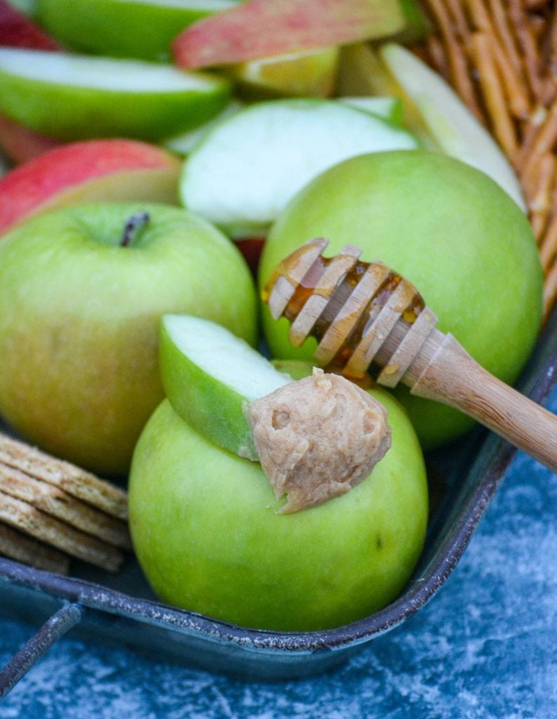 a scoop of healthy peanut butter apple dip shown on a green apple slice resting on top of a whole green apple with a honey covered wooden dipper behind it