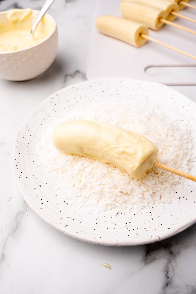 a white chocolate coated banana pop on a plate with coconut flakes