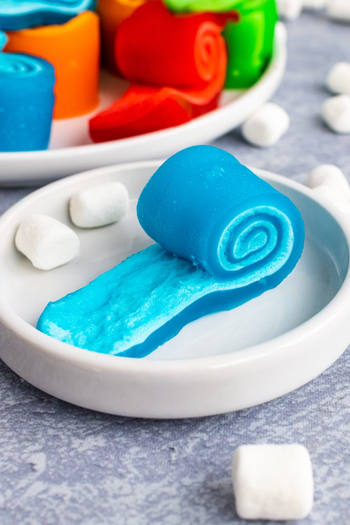a blue jello marshmallow pinwheel on a small white plate surrounded by mini marshmallows