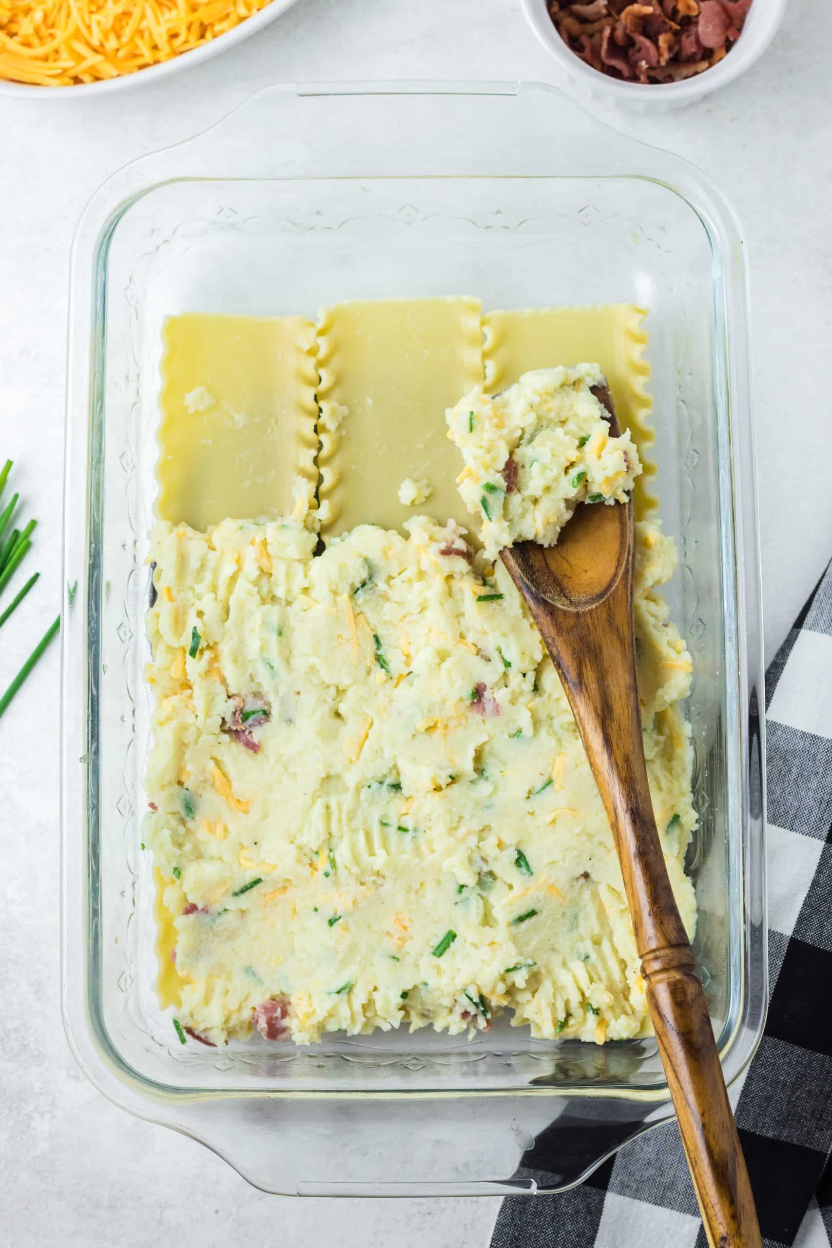 bacon cheddar mashed potatoes being spread over cooked lasagna noodles with a wooden spoon in a glass baking dish