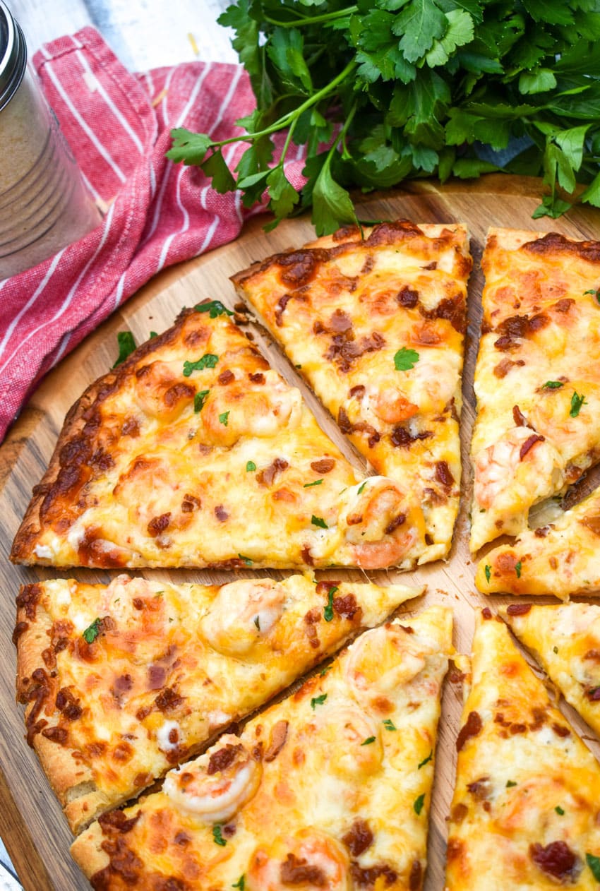 a sliced garlic shrimp alfredo pizza on a wooden pizza peel with fresh herbs in the background