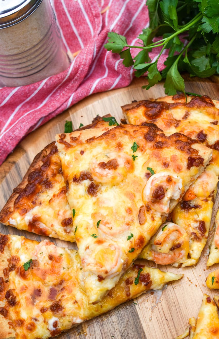 a sliced garlic shrimp alfredo pizza on a wooden pizza peel with fresh herbs in the background