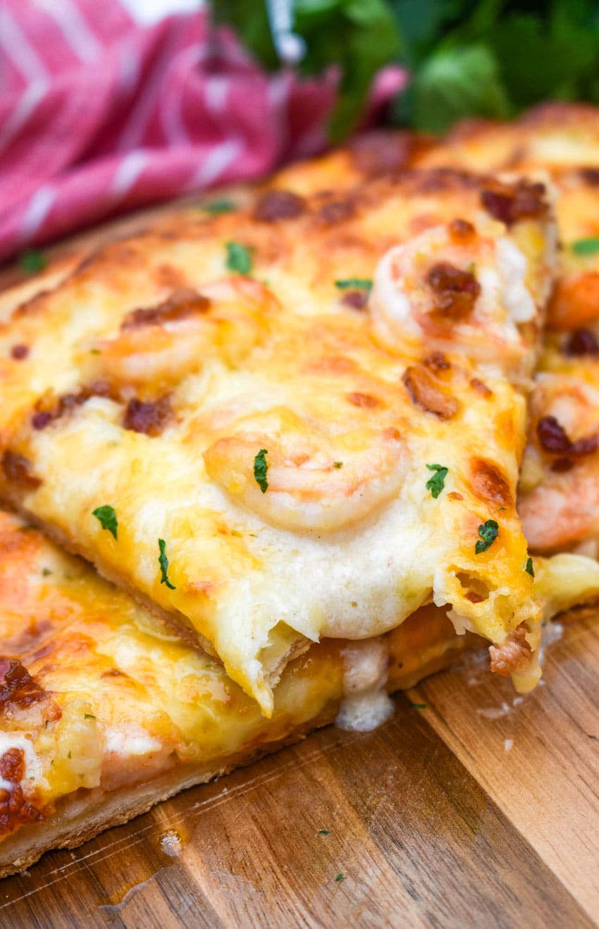 a sliced garlic shrimp alfredo pizza stacked on a wooden pizza peel with fresh herbs in the background