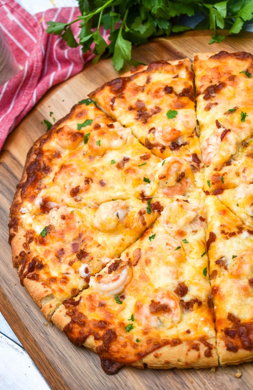 a sliced garlic shrimp alfredo pizza on a wooden pizza peel with fresh herbs in the background