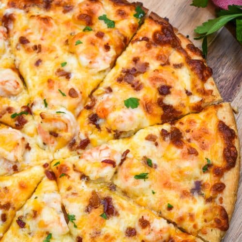 a sliced garlic shrimp alfredo pizza on a wooden pizza peel with fresh herbs in the background