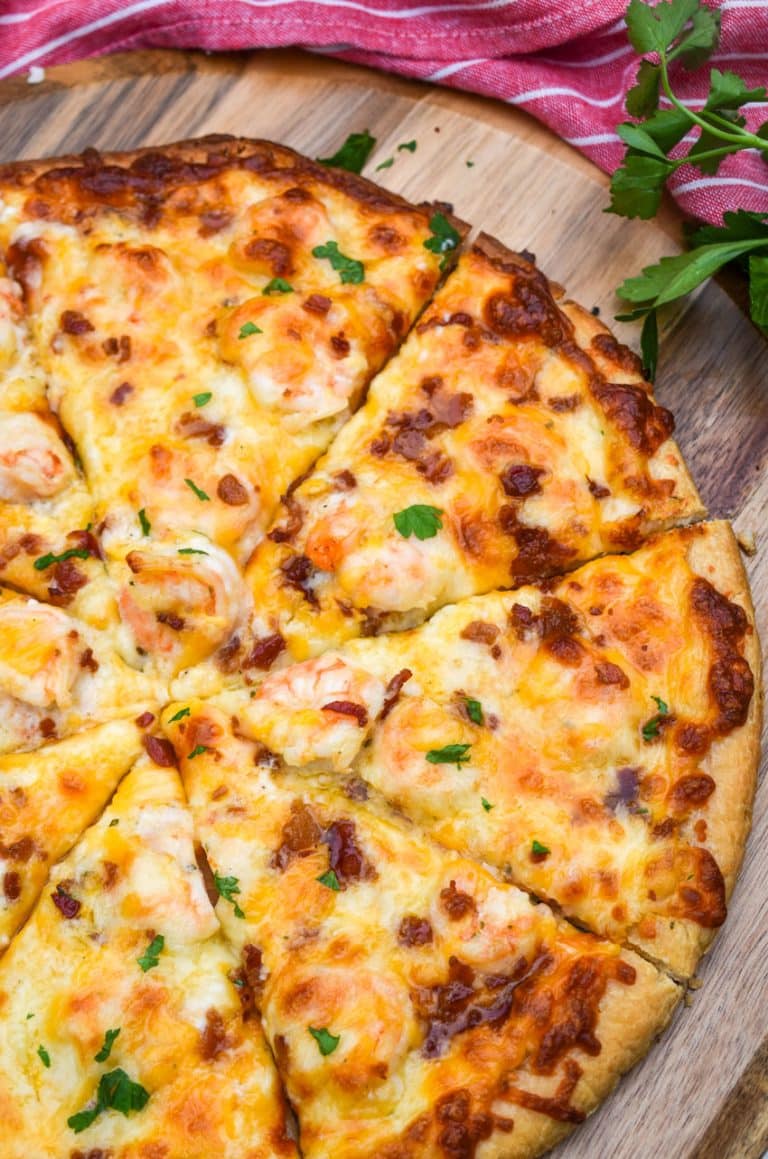 a sliced garlic shrimp alfredo pizza on a wooden pizza peel with fresh herbs in the background