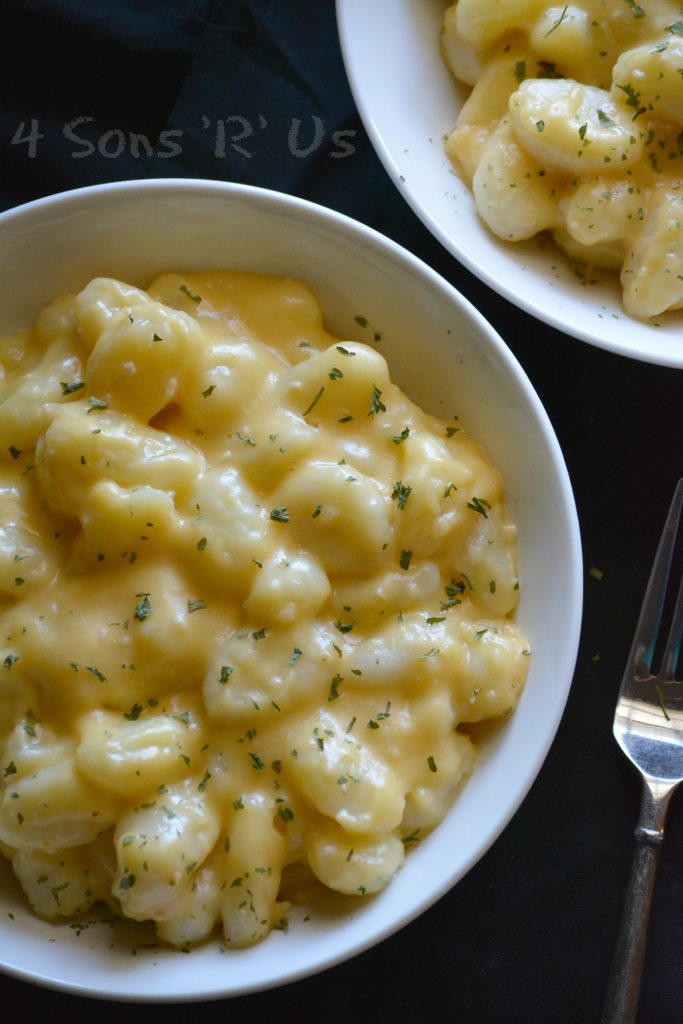 Fluffy potato gnocchi are bathed in a thick creamy cheese sauce all piled high in a white bowl on a black back ground with a fork to the side and a sprinkle or parsley leaves on top.