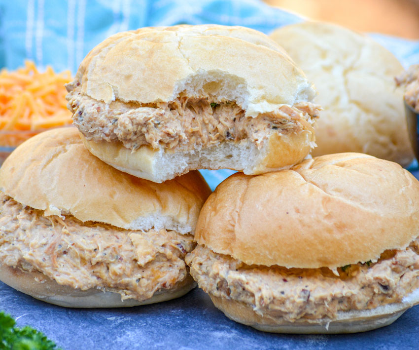 a crockpot crack chicken sandwich shown with a bite taken out of it and sitting on top of two other prepared crack chicken sandwiches