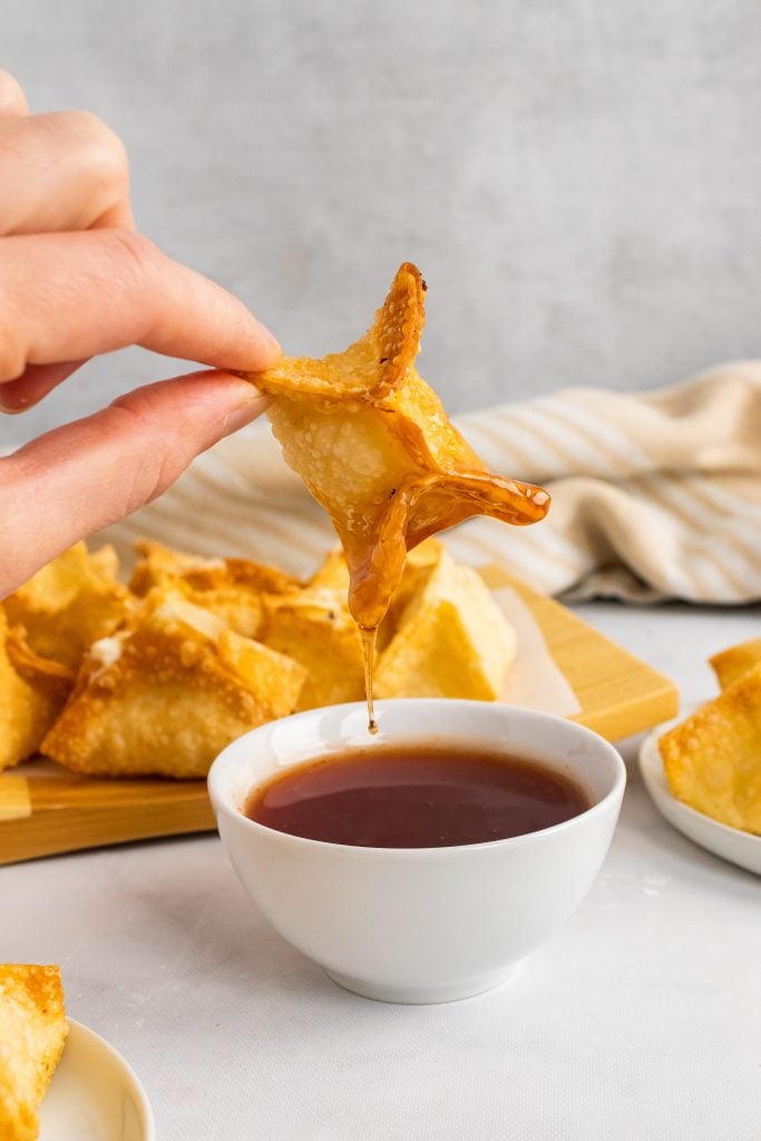 a hand holding up a crab rangoon dipped in sauce with drops of sauce falling in a bowl below it