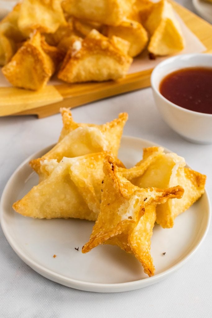 crispy fried crab rangoon on a small white plate with a bowl of dipping sauce in the background