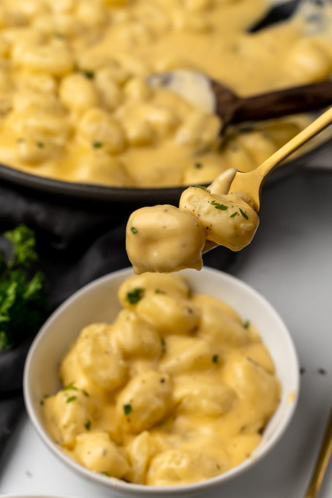 a gold fork holding up cheesy gnocchi above a white bowl filled with gnocchi mac and cheese