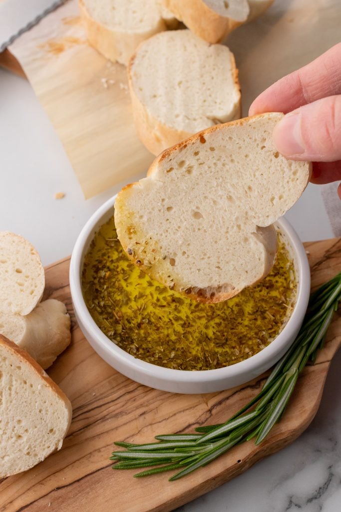 a hand dipping a slice of 30 minute french bread into a shallow white bowl filled with dipping oil