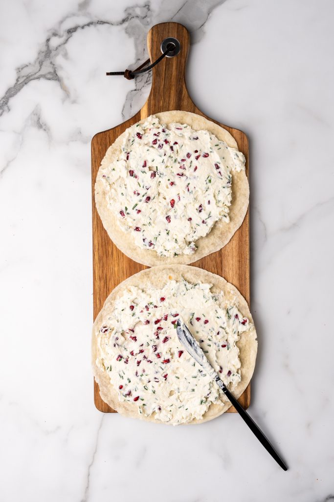 pomegranate feta and chive filling spread on two large flour tortillas sitting on a wooden cutting board