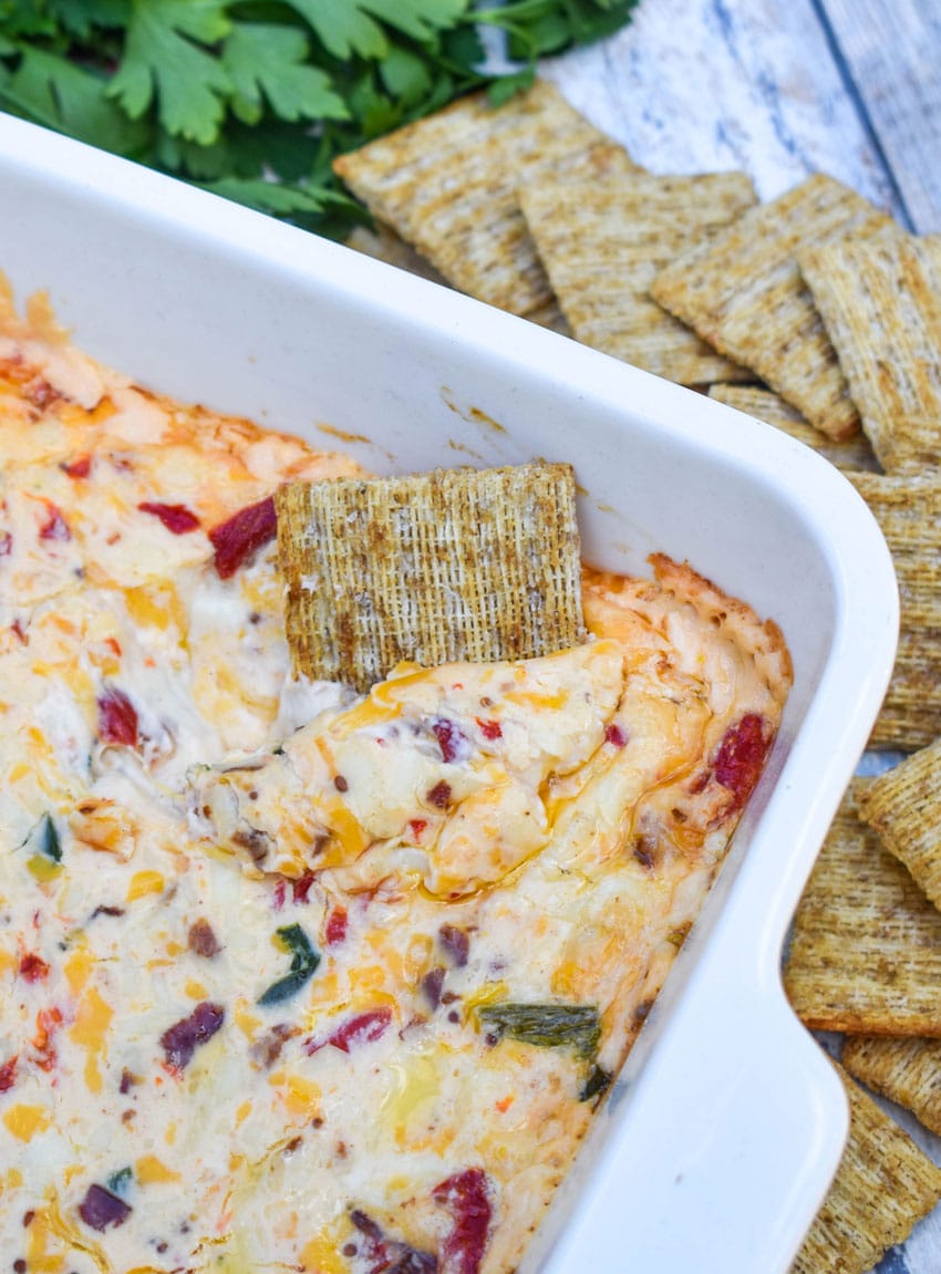 a cracker scooping hot pimento cheese dip out of a white baking dish