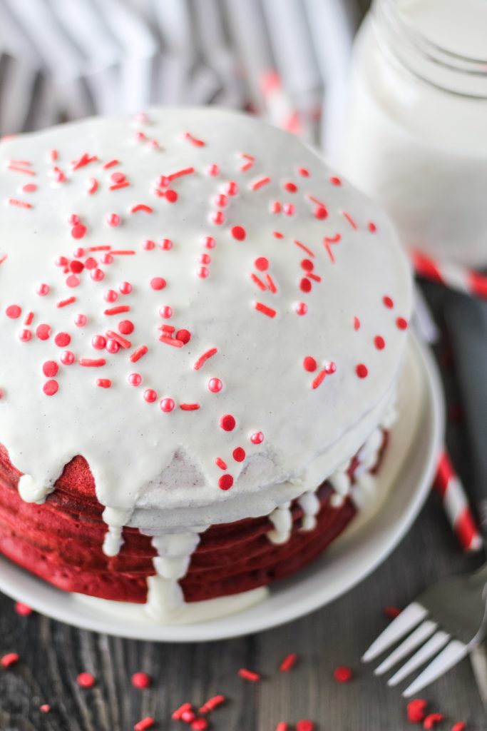 red velvet pancakes topped with cream cheese glaze stacked on a white plate