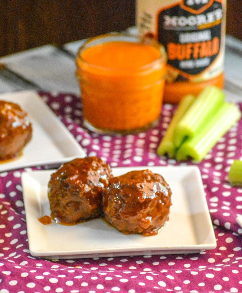 two Crockpot Honey Buffalo Meatballs on a small square white plate with a glass of wing sauce and cut celery sticks in the background