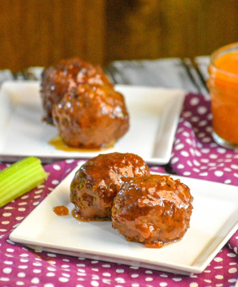 two Crockpot Honey Buffalo Meatballs on a small square white plate with a glass of wing sauce and cut celery sticks in the background
