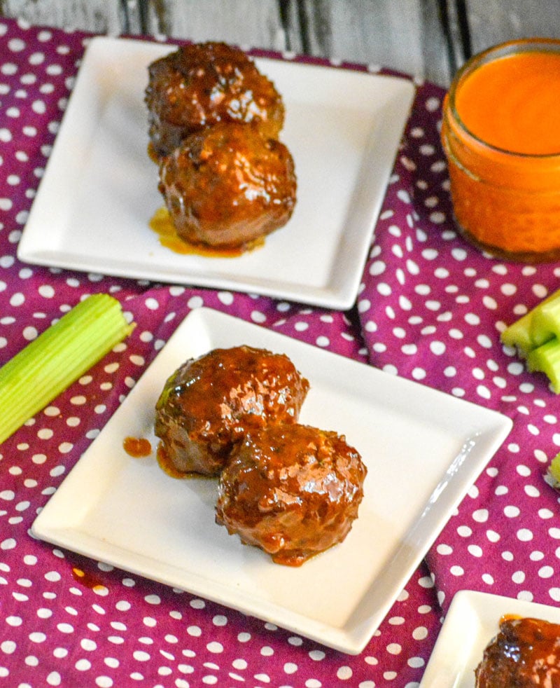 two saucey Honey Buffalo Meatballs each shown on a small square white plate with a glass of wing sauce and cut celery sticks in the background