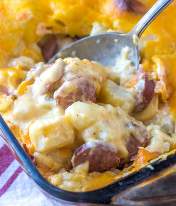 a silver spoon digging into a baking dish filled with cheesy potato and smoked sausage casserole