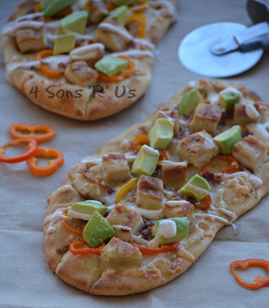 naan bread on parchment paper has been topped with chunks of cooked chicken, avocados, and sweet pepper rings with a chipotle ranch drizzle. Extra sweet pepper slices and a pizza cutter are shown in the background.