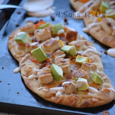 naan bread on a sheet pan has been topped with chunks of cooked chicken, avocados, and sweet pepper rings with a chipotle ranch drizzle. A spoon with chipotle ranch is seen in the background.