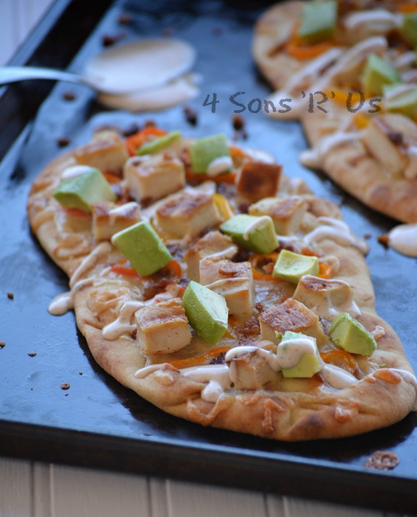 naan bread on a sheet pan has been topped with chunks of cooked chicken, avocados, and sweet pepper rings with a chipotle ranch drizzle. A spoon with chipotle ranch is seen in the background.