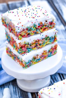 a stack of frosting topped fruity pebbles cereal bars on a small white dessert stand