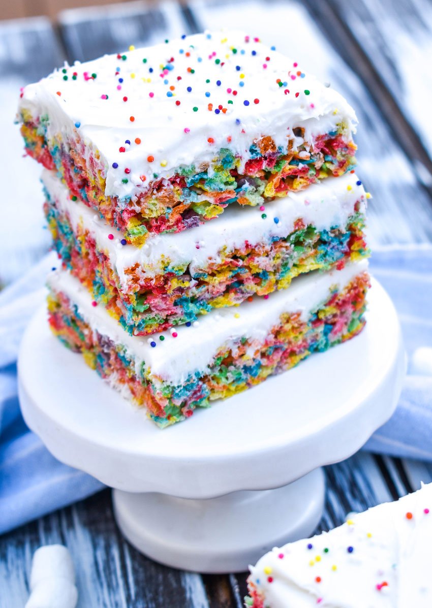 a stack of frosting topped fruity pebbles cereal bars on a small white dessert stand