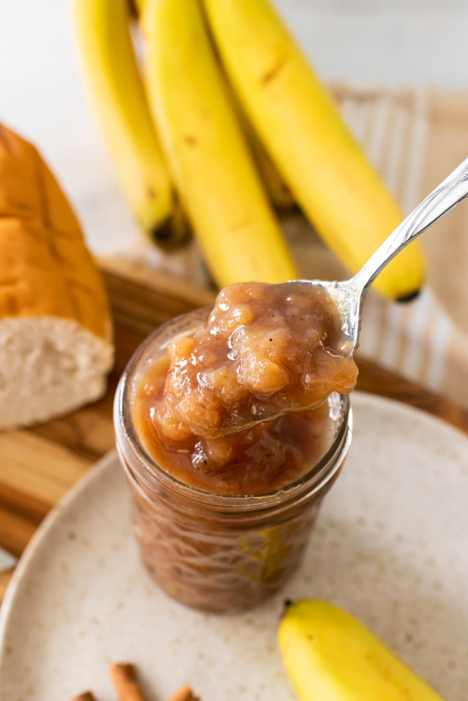 a silver spoon shown lifting a scoop of banana rum jam out of a glass jar