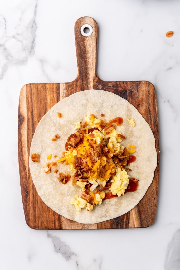 cheesy bacon topped eggs and hashbrown patty on a large flour tortilla on a wooden cutting board