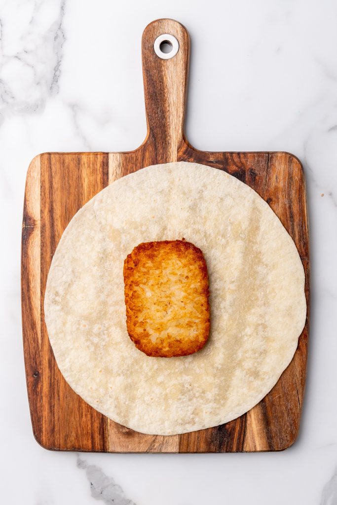 a crispy cooked hashbrown patty in the center of a large flour tortilla on a wooden cutting board